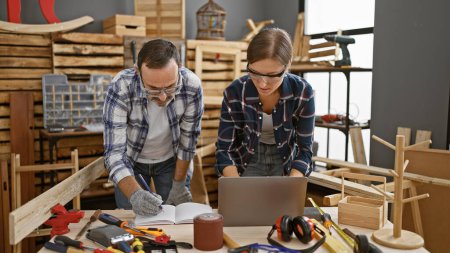 Photo for Two hardworking carpenters, man and woman, collaborating online using laptop, seriously taking notes at their indoor carpentry business worksite amidst timber and woodwork equipment - Royalty Free Image