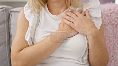 Foto de Young blonde woman sitting on sofa with hands on heart at home - Imagen libre de derechos