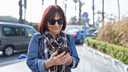 Foto de Una mujer hispana feliz y madura con gafas de sol utiliza un teléfono inteligente en una calle urbana soleada con coches y árboles en el fondo. - Imagen libre de derechos