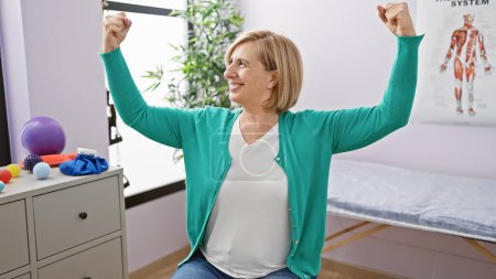 Photo for A smiling, middle-aged blonde woman flexing her muscles in a rehab clinic room, showcasing strength and positivity. - Royalty Free Image