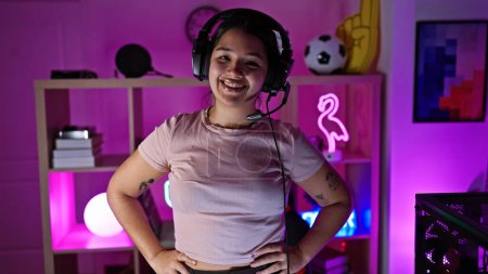Foto de Mujer hispana con auriculares sonriendo en una sala de juegos iluminada por neón en casa, proyectando juventud y tecnología. - Imagen libre de derechos
