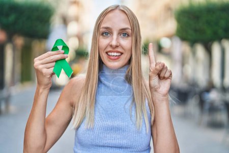 Téléchargez les photos : Jeune femme caucasienne tenant le soutien ruban vert souriant avec une idée ou une question pointant du doigt avec un visage heureux, numéro un - en image libre de droit