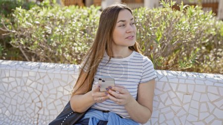 Eine nachdenkliche junge Frau mit langen Haaren hält in einem sonnigen Park ein Smartphone in der Hand und strahlt inmitten der Natur heitere Schönheit aus..