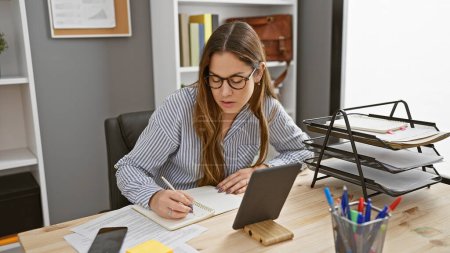 Foto de Mujer morena enfocada escribiendo en cuaderno en su escritorio de oficina, en interiores con tableta digital - Imagen libre de derechos