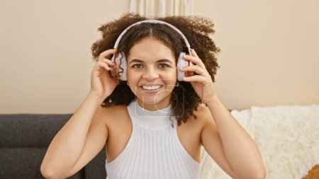 Foto de Sonriente joven hispana con el pelo rizado usando auriculares, disfrutando de la música en casa, sentada en un sofá. - Imagen libre de derechos