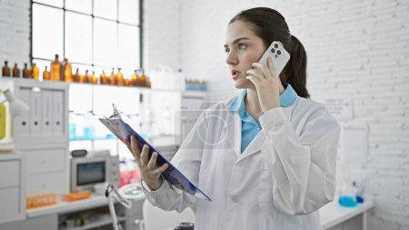 Photo for A concentrated young woman wearing a lab coat talks on the phone in a modern laboratory while holding a clipboard. - Royalty Free Image