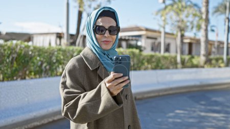 A mature hijab-wearing woman stands outside using a smartphone, with sunglasses and a coat, in a sunny, urban setting.