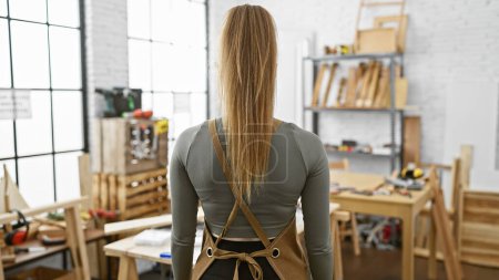 Photo for Back view of a young woman with blonde hair standing in a well-lit carpentry workshop, portraying a craftsman atmosphere. - Royalty Free Image