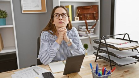 Photo for Pensive woman with glasses and long hair contemplating at her office desk surrounded by documents and a laptop. - Royalty Free Image