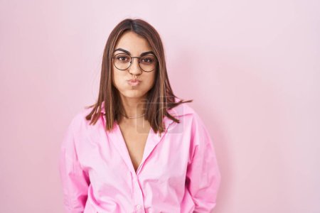 Foto de Mujer hispana joven con gafas de pie sobre fondo rosa hinchando mejillas con cara divertida. boca hinchada de aire, expresión loca. - Imagen libre de derechos