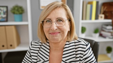 Téléchargez les photos : Une femme d'âge moyen souriante avec des lunettes dans un blazer rayé se trouve dans un bureau moderne - en image libre de droit