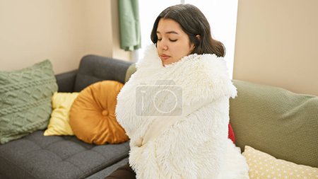 Foto de Una joven hispana envuelta en una acogedora manta, descansa pacíficamente en un sofá en una sala de estar bien decorada. - Imagen libre de derechos