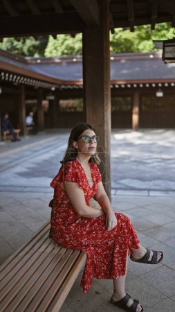 Téléchargez les photos : Belle femme hispanique aux lunettes fourrées dans la solitude tranquille assis sur un banc au temple Meiji de Tokyo, profitant de tout le vert de l'été au milieu de l'agitation de la culture japonaise. - en image libre de droit