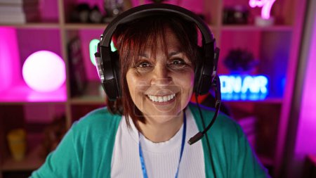 Une femme hispanique mature souriante portant un casque dans une salle de jeux colorée la nuit