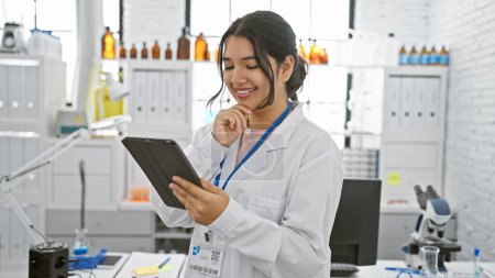 Eine lächelnde junge hispanische Frau im Laborkittel benutzt in einem modernen Labor eine Tablette, die für medizinische Professionalität und Technologie steht..