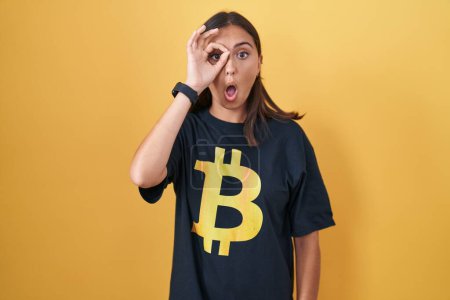 Téléchargez les photos : Jeune femme hispanique portant Bitcoin t-shirt faire un geste correct choqué par le visage surpris, oeil regardant à travers les doigts. expression incrédule. - en image libre de droit