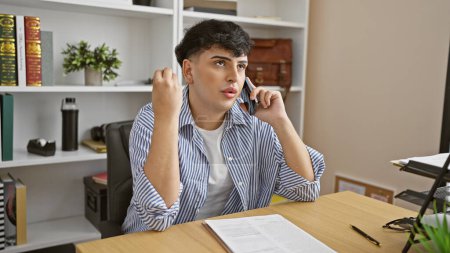 Photo for A young man in a striped shirt is on a phone call at an organized office desk with bookshelves in the background. - Royalty Free Image