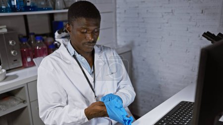 Foto de Científico africano en bata de laboratorio poniéndose guantes azules en laboratorio, enfocado en su tarea - Imagen libre de derechos