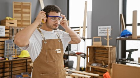 A bearded craftsman adjusting safety goggles in a well-organized carpentry studio with tools and wood.