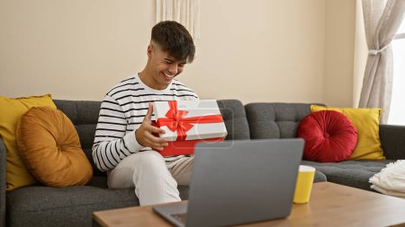 Téléchargez les photos : Surpris jeune homme hispanique assis à la maison sur le canapé, déballer cadeau surprise tout en parlant joyeusement et en souriant lors d'un appel vidéo en ligne. - en image libre de droit
