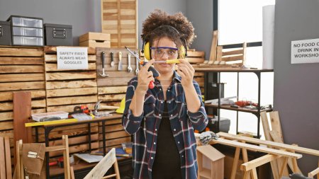 Photo for Young hispanic woman with curly hair wearing safety goggles and ear protection in a carpentry workshop - Royalty Free Image