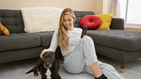 A smiling young woman in casual attire enjoys time indoors with her black labrador, while holding a smartphone in a cozy living room setting.