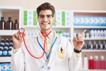 Téléchargez les photos : Jeune homme hispanique travaillant à la pharmacie pharmacie en utilisant stéthoscope souriant heureux pointant avec la main et le doigt vers le côté - en image libre de droit