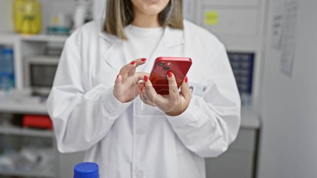 Téléchargez les photos : Jeune femme en blouse de laboratoire à l'aide d'un smartphone en laboratoire - en image libre de droit