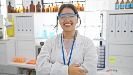 Foto de Retrato de una joven hispana sonriente con los brazos cruzados, de pie en un entorno de laboratorio. - Imagen libre de derechos