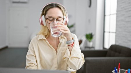 Téléchargez les photos : Jeune femme blonde travailleuse d'affaires utilisant un ordinateur portable et des écouteurs d'eau potable au bureau - en image libre de droit
