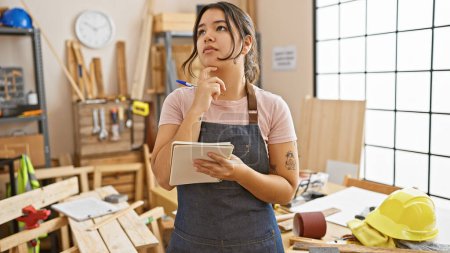 Photo for Thoughtful hispanic woman with tablet pondering in a carpentry workshop surrounded by wood, tools, and safety helmet. - Royalty Free Image