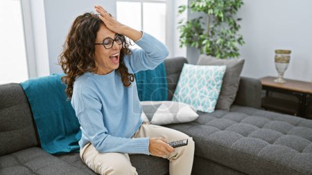 Photo for Hispanic woman laughing in living room holding remote, expressing joy and comfort on a cozy sofa indoors. - Royalty Free Image