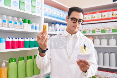 Foto de Joven hombre hispano farmacéutico sosteniendo pastillas botella lectura prescripción en farmacia - Imagen libre de derechos