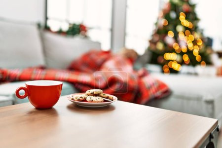 Cozy christmas scene with festive mug, cookies, blanket, and tree lights in blurred background.