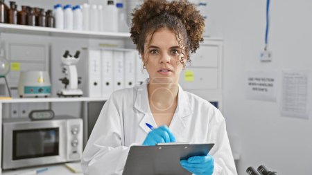 A young hispanic woman with curly hair, wearing a lab coat and gloves, takes notes in a laboratory setting.