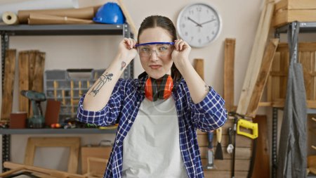 Photo for Caucasian woman with safety goggles in a carpentry workshop surrounded by wood and tools - Royalty Free Image