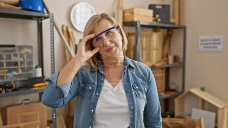 Foto de Una mujer rubia de mediana edad con gafas de seguridad está de pie con confianza en un taller de carpintería. - Imagen libre de derechos