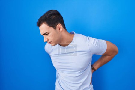 Téléchargez les photos : Jeune homme hispanique debout sur fond bleu souffrant de maux de dos, touchant le dos avec la main, douleurs musculaires - en image libre de droit
