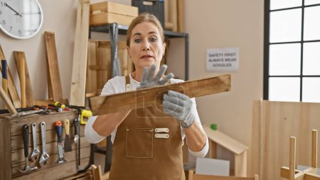 Téléchargez les photos : Femme mûre examine la qualité du bois dans un atelier de menuiserie, entouré d'outils et d'établi. - en image libre de droit