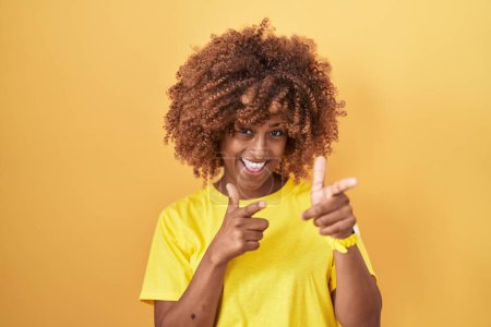 Foto de Young hispanic woman with curly hair standing over yellow background pointing fingers to camera with happy and funny face. good energy and vibes. - Imagen libre de derechos