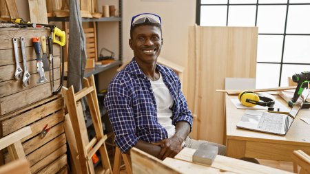 Foto de Un hombre afroamericano sonríe sentado con confianza en su taller de carpintería lleno de varias herramientas y equipos. - Imagen libre de derechos