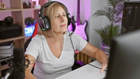Focused middle-aged woman with headphones in a gaming room using a microphone and computer at night.