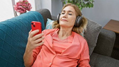 Téléchargez les photos : Femme blonde mature se détend à la maison sur le canapé en utilisant un smartphone et un casque, représentant les loisirs et la technologie. - en image libre de droit