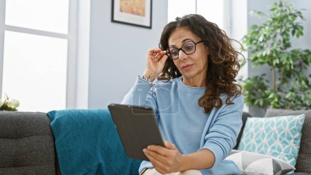 Foto de Una mujer hispana de mediana edad con cabello rizado lee desde una tableta en su acogedora y moderna sala de estar. - Imagen libre de derechos