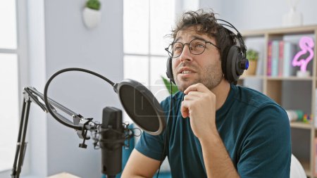 Téléchargez les photos : Bel homme hispanique avec barbe et écouteurs contemplant lors de l'enregistrement dans un studio de radio. - en image libre de droit