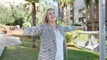 Téléchargez les photos : Femme blonde mature étirant les bras avec joie à l'extérieur dans un parc urbain verdoyant. - en image libre de droit