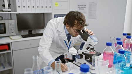 Foto de Científico joven serio, un rubio barbudo, meticulosamente tomando notas en la mesa de laboratorio, absorto en la investigación médica bajo el microscopio en un bullicioso laboratorio de ciencias. - Imagen libre de derechos