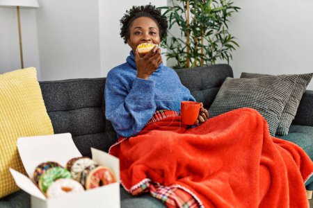 Téléchargez les photos : Femme afro-américaine prenant le petit déjeuner assis sur le canapé à la maison - en image libre de droit