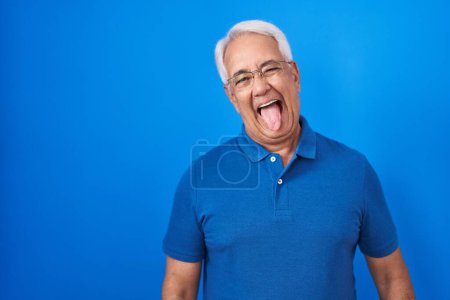 Téléchargez les photos : Middle age man with grey hair standing over blue background sticking tongue out happy with funny expression. emotion concept. - en image libre de droit