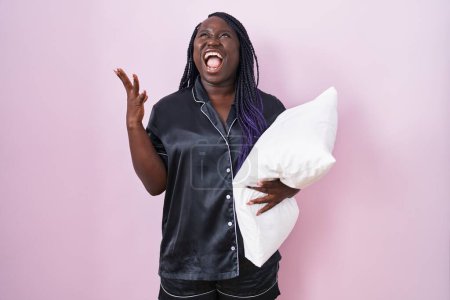 Foto de Mujer africana joven con pijama abrazando almohada loca y loca gritando y gritando con expresión agresiva y los brazos levantados. concepto de frustración. - Imagen libre de derechos